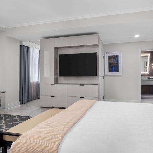 A modern hotel room features a bed, TV, armchair, and a bathroom with illuminated mirrors.