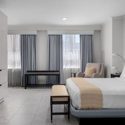 A modern hotel room with a bed, TV, armchair, and curtains.