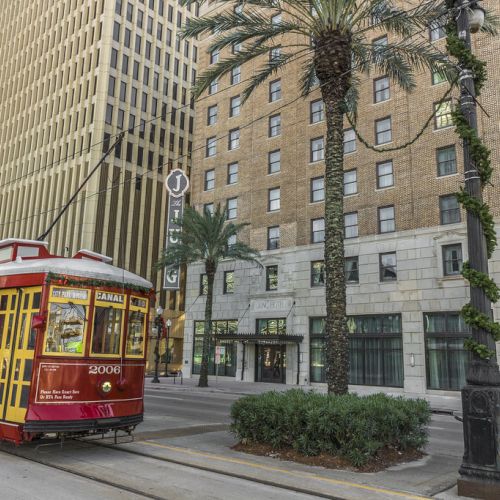 A red streetcar travels past a city street flanked by palm trees and tall buildings on a sunny day.