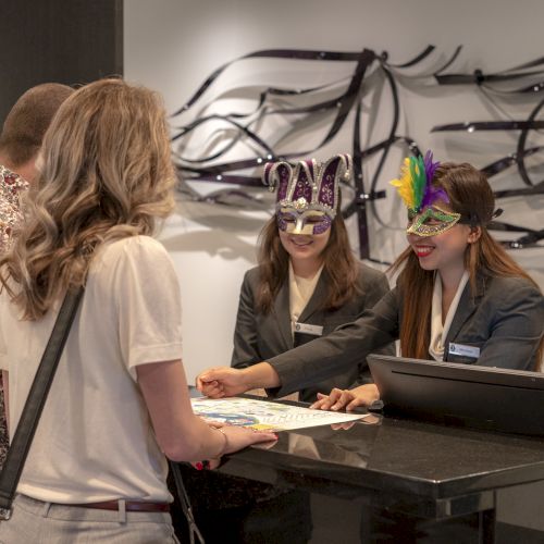 Two people are at a reception desk, assisted by staff wearing colorful masks, discussing something on a document.