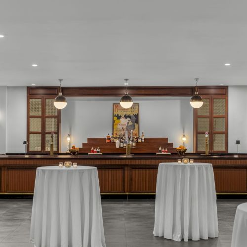 The image shows an elegant bar with round tables covered in white cloth, lit by pendant lights, creating a sophisticated atmosphere.