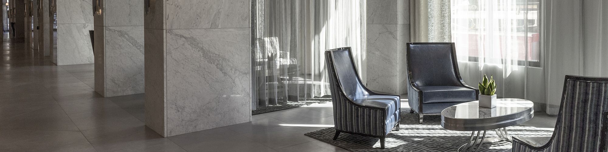 A modern lobby with marble walls, sleek chairs, a coffee table, decorative lighting, and sheer curtains filtering sunlight.