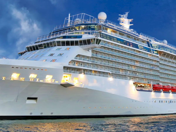 A massive white cruise ship docked at port, multiple decks with windows, balconies, and bright lights against a blue sky, peaceful water.