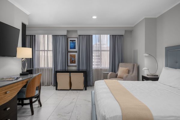 A modern hotel room with a bed, desk, chair, TV, and large windows with curtains, featuring neutral tones and minimalistic decor.