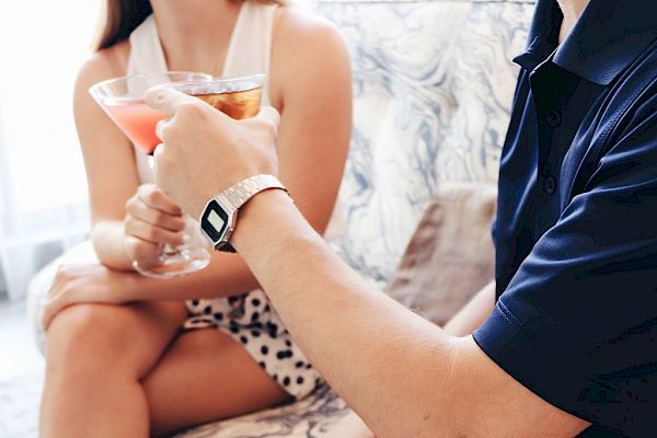 Two people sitting on a couch, holding cocktails, one with a pink drink and the other with an amber drink.