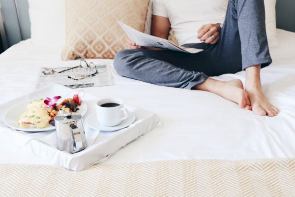 A person relaxes on a bed with a breakfast tray holding coffee, a meal, and a magazine, creating a cozy morning scene.