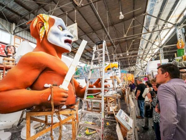 An artist wearing an orange mask and costume sculpts a large white mask with a sword in a busy workshop, surrounded by ladders and onlookers.
