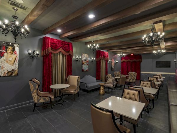 A chic lounge with dark walls, red drapes, chandeliers, and tufted seating; upholstered chairs, small tables, and a cozy, upscale vibe.