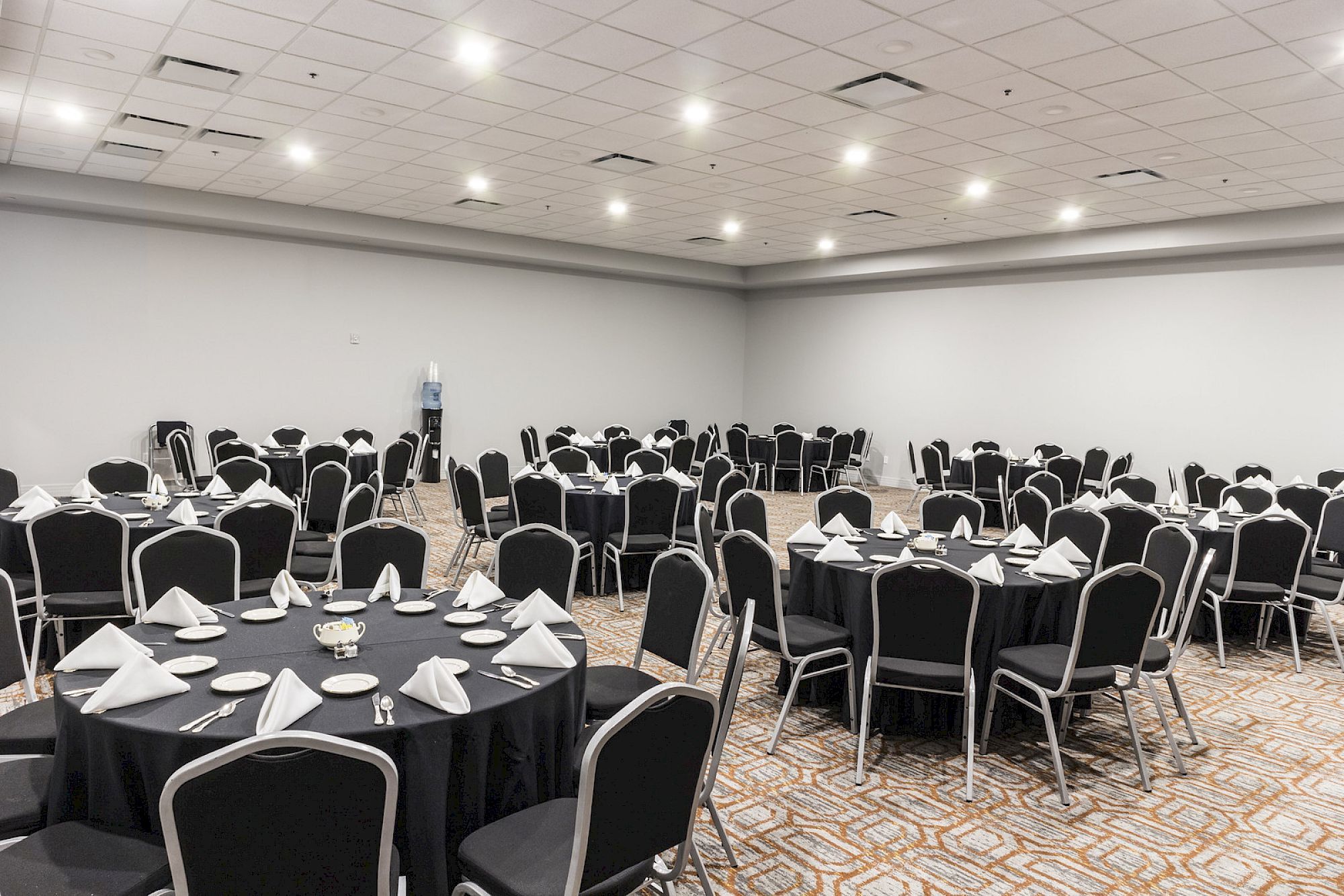 A conference banquet hall with round tables, black chairs, white napkins, and place settings ready for an event.