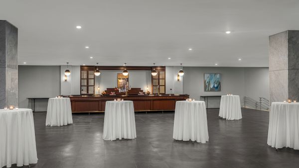 A dimly lit bar area with round tables draped in white tablecloths, candles on each table, and modern hanging lights.
