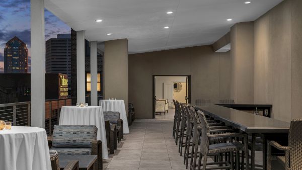 Outdoor terrace with round and rectangular tables, wicker chairs, and a skyline view, likely in a city setting, during the evening.