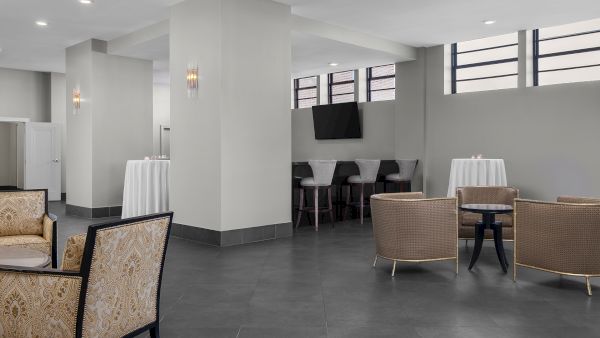 A spacious lounge with elegant seating, high tables, bar stools, and a mounted TV. Neutral tones and modern decor create a cozy ambiance.